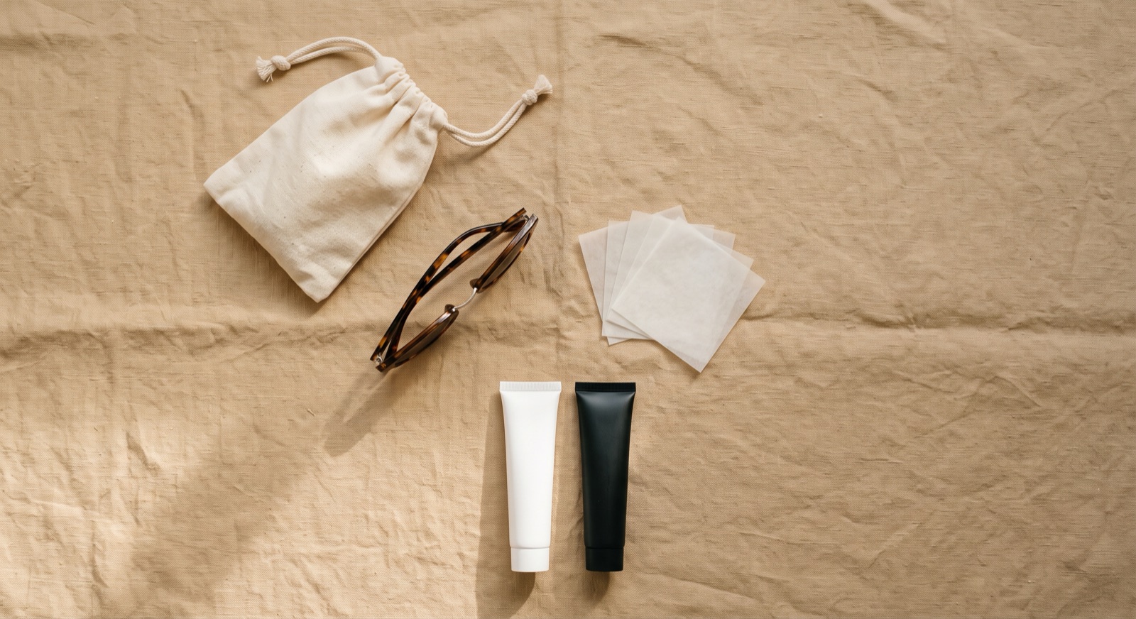 A flat lay on sand linen: a small pouch, sunglasses, blotting papers and two plain cosmetic tubes